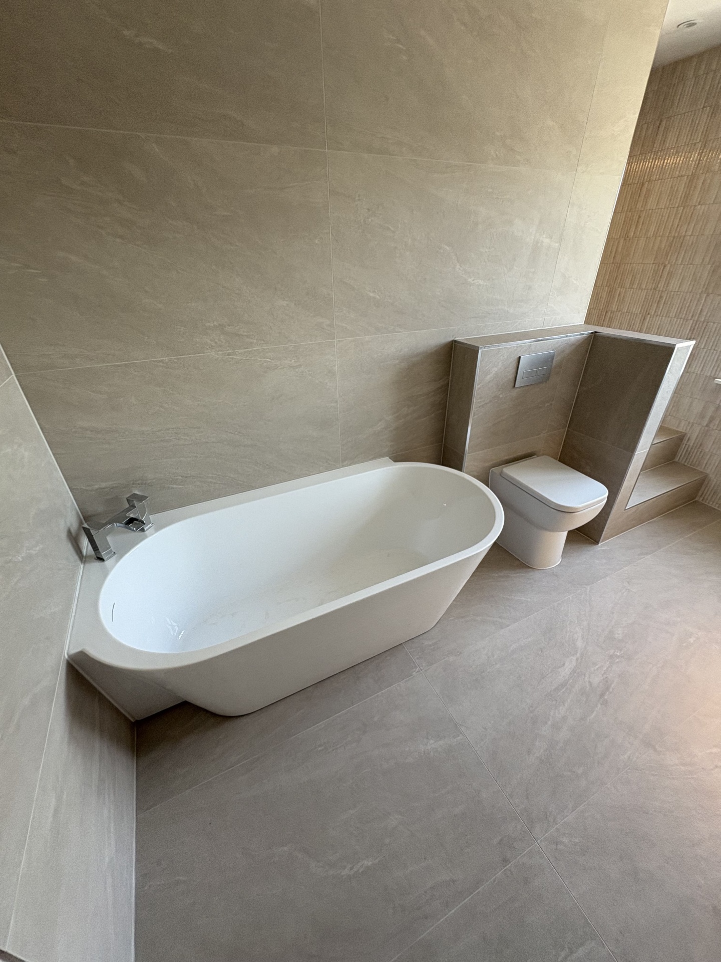 Completed modern bathroom featuring a freestanding bath, wall-mounted toilet and large beige wall tiles