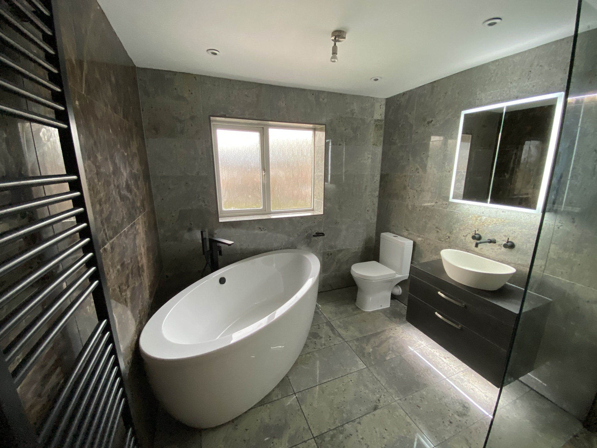 Completed grey-tiled bathroom with freestanding bath, illuminated mirror and walk-in shower